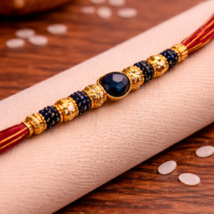 Close-up of designer rakhi with navy blue gemstone centerpiece, gold beads, and intricate detailing placed on beige fabric.