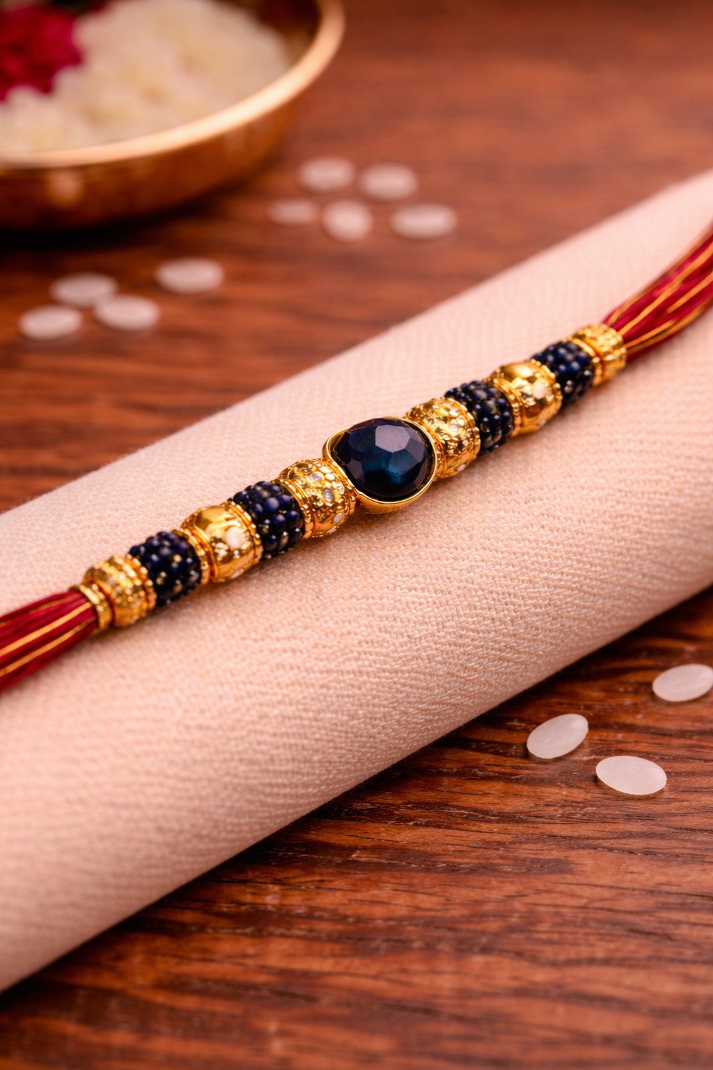 Close-up of designer rakhi with navy blue gemstone centerpiece, gold beads, and intricate detailing placed on beige fabric.