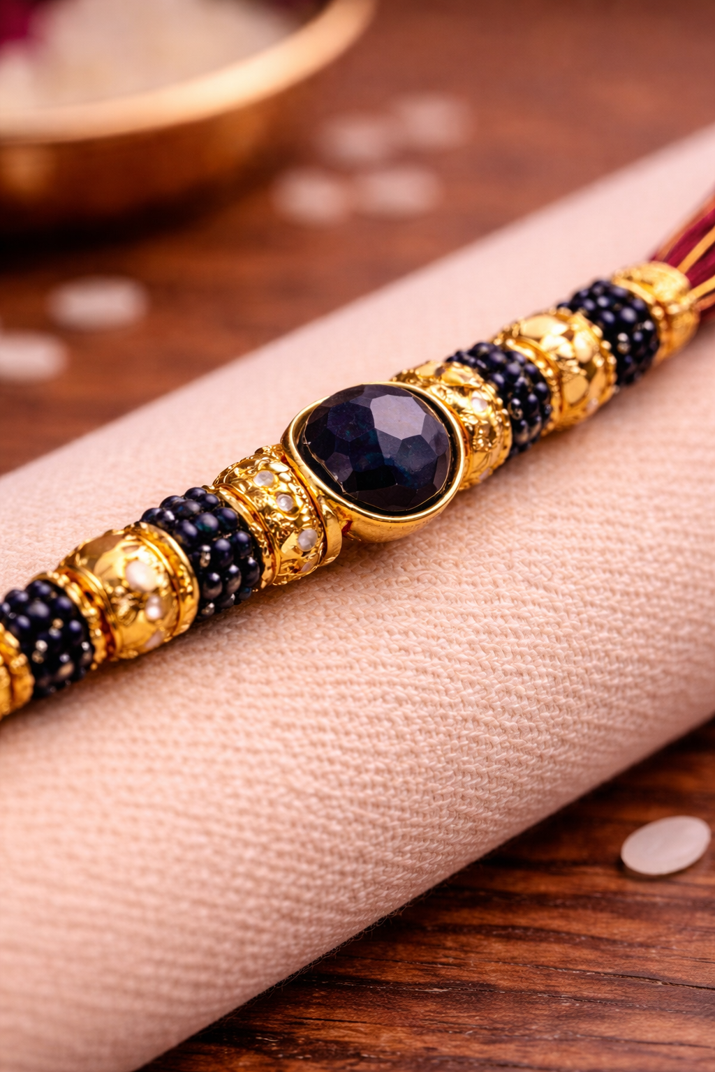 Close-up of designer rakhi with navy blue gemstone centerpiece, gold beads, and intricate detailing placed on beige fabric.